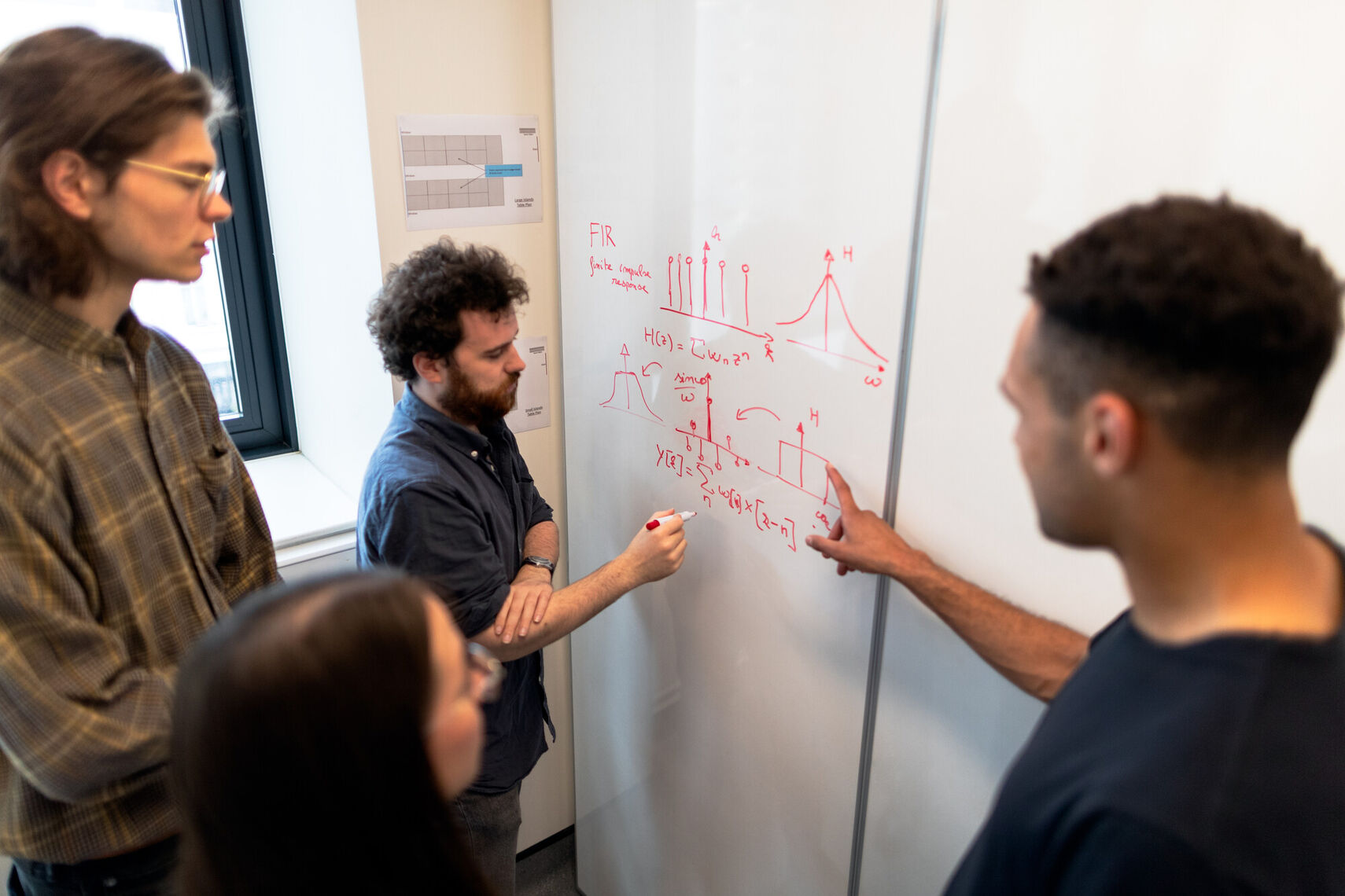 One person writes some calculations on a white board while three others are looking and one points. 