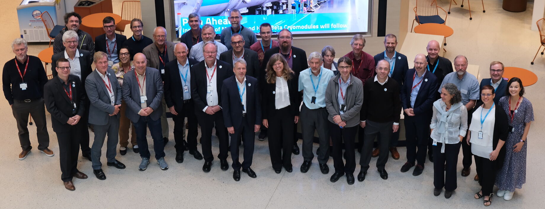 25 members of the LENS meeting stand in front of a screen in the atrium of the ESS offices